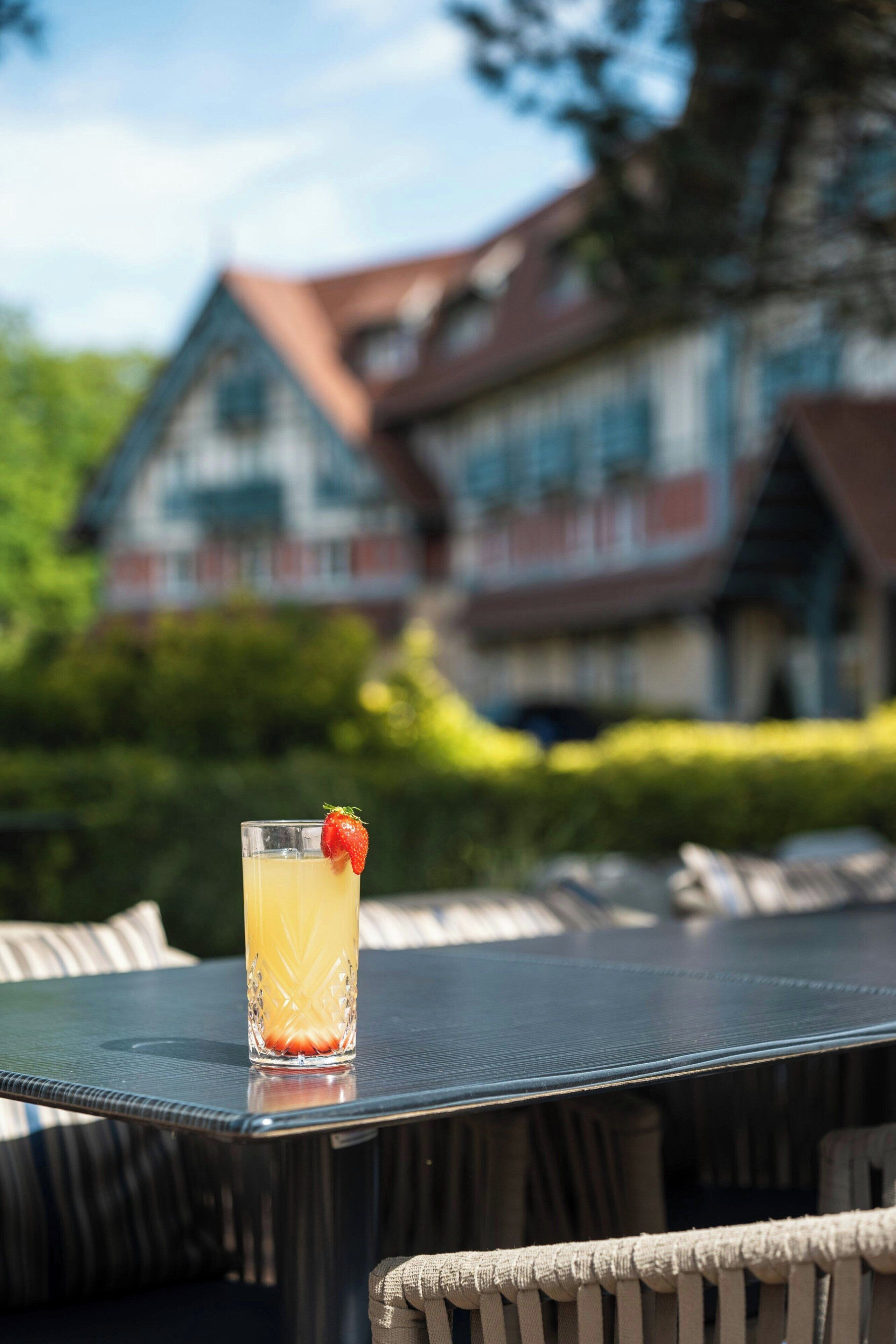 Un verre contenant une boisson, décoré d’une fraise, est posé sur une table à l’extérieur, avec un bâtiment en arrière-plan.