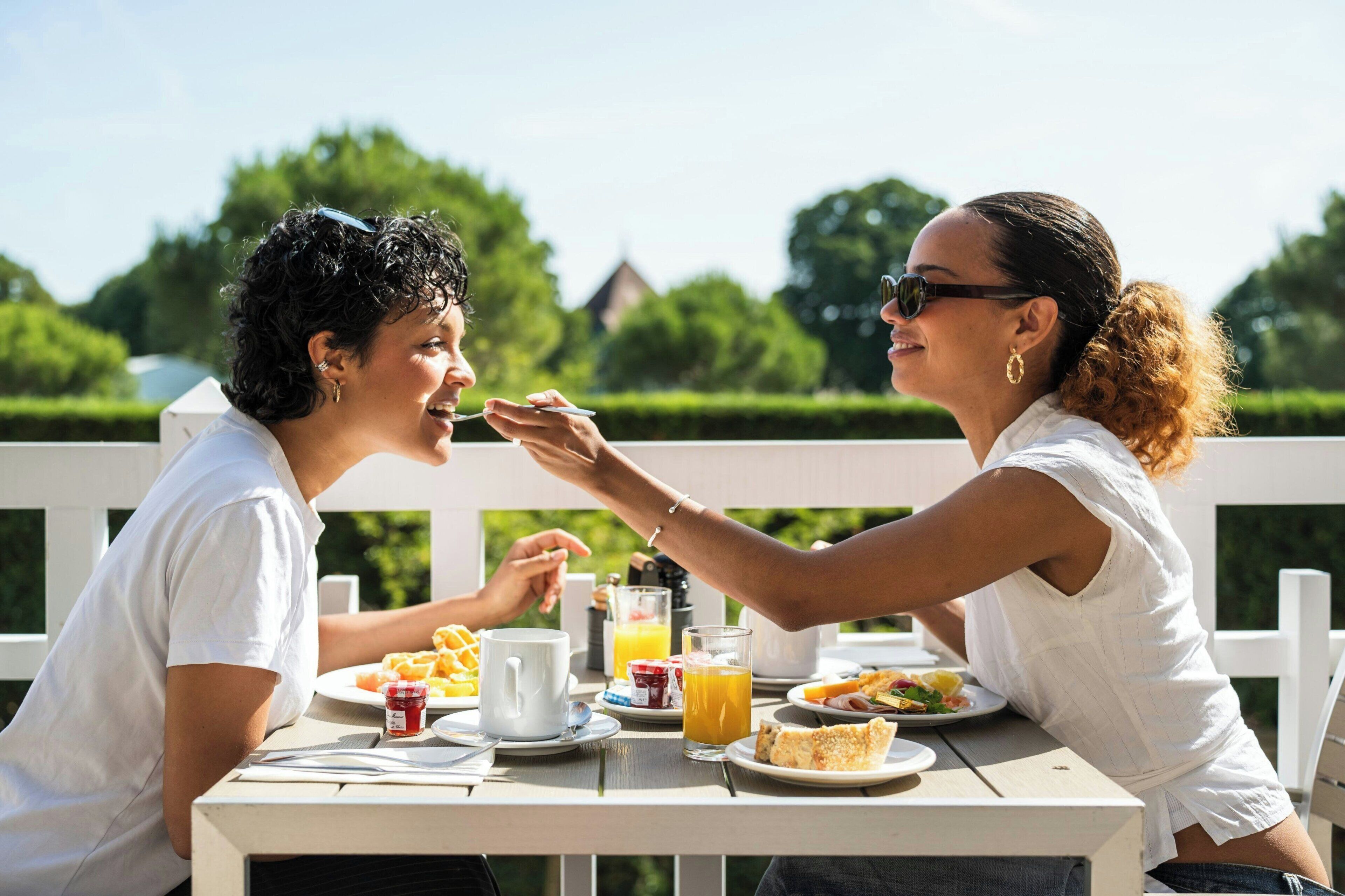 Dos personas sentadas en una mesa al aire libre con alimentos para el desayuno, como tostadas, fruta y jugo.