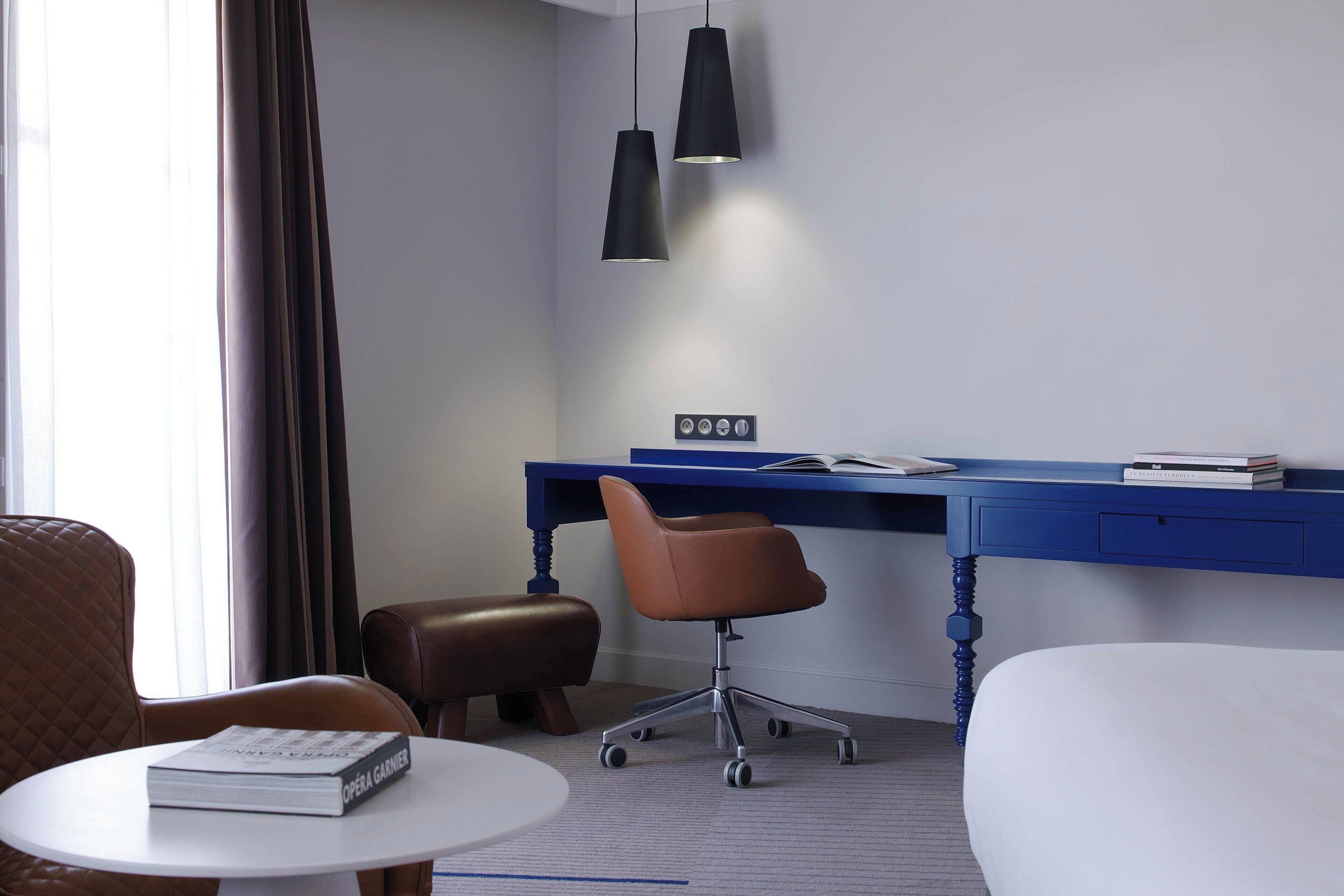 Ein Hotelzimmer mit blauem Schreibtisch, braunem Ledersessel, weißem Bett und kleinem runden Tisch mit einem Buch.
