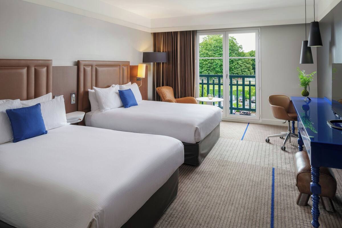 A hotel room with two beds, a desk, a chair, and a balcony with a view of greenery.