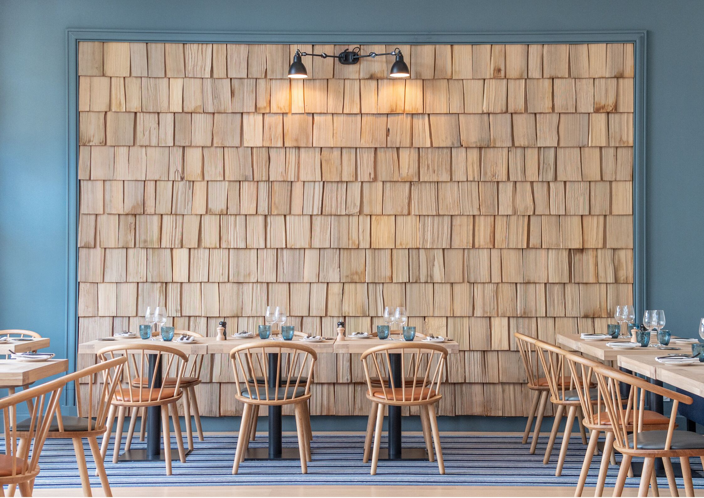 A restaurant interior with a wooden shingle wall, round tables, and wooden chairs.