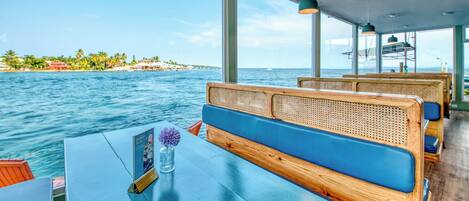 A boat with wooden benches and tables, large windows offering a view of the sea and a distant shoreline.