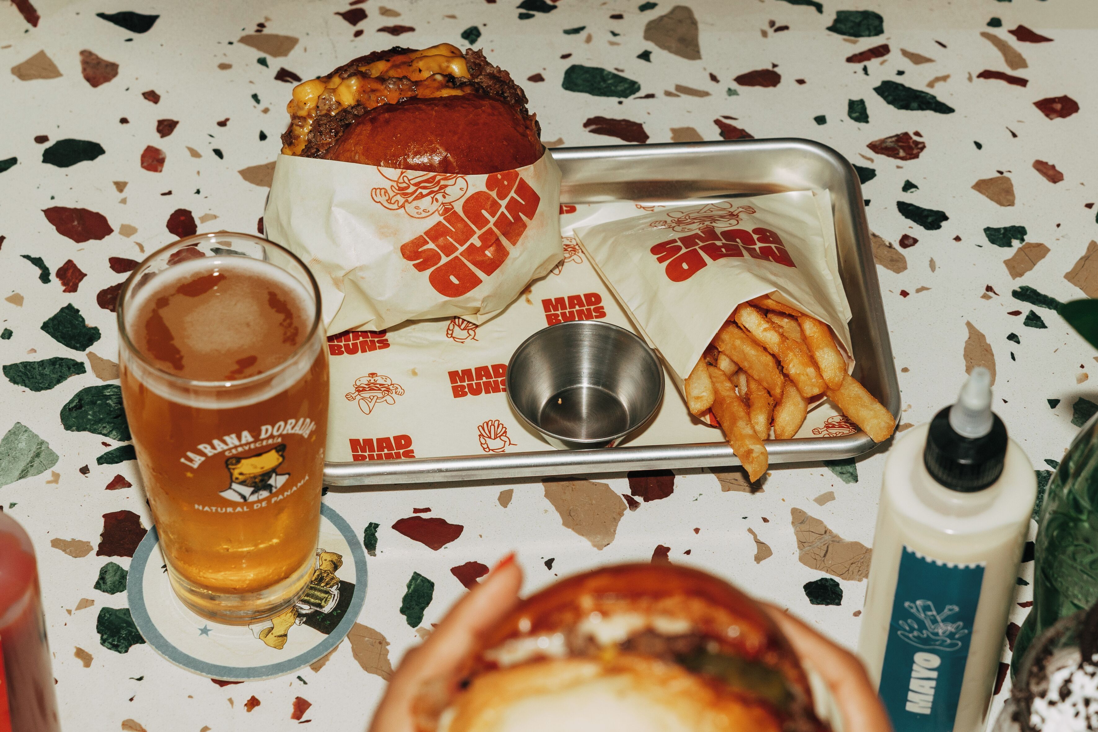 A meal tray with a burger, fries, and a drink, along with a bottle of sauce and a glass of beer.