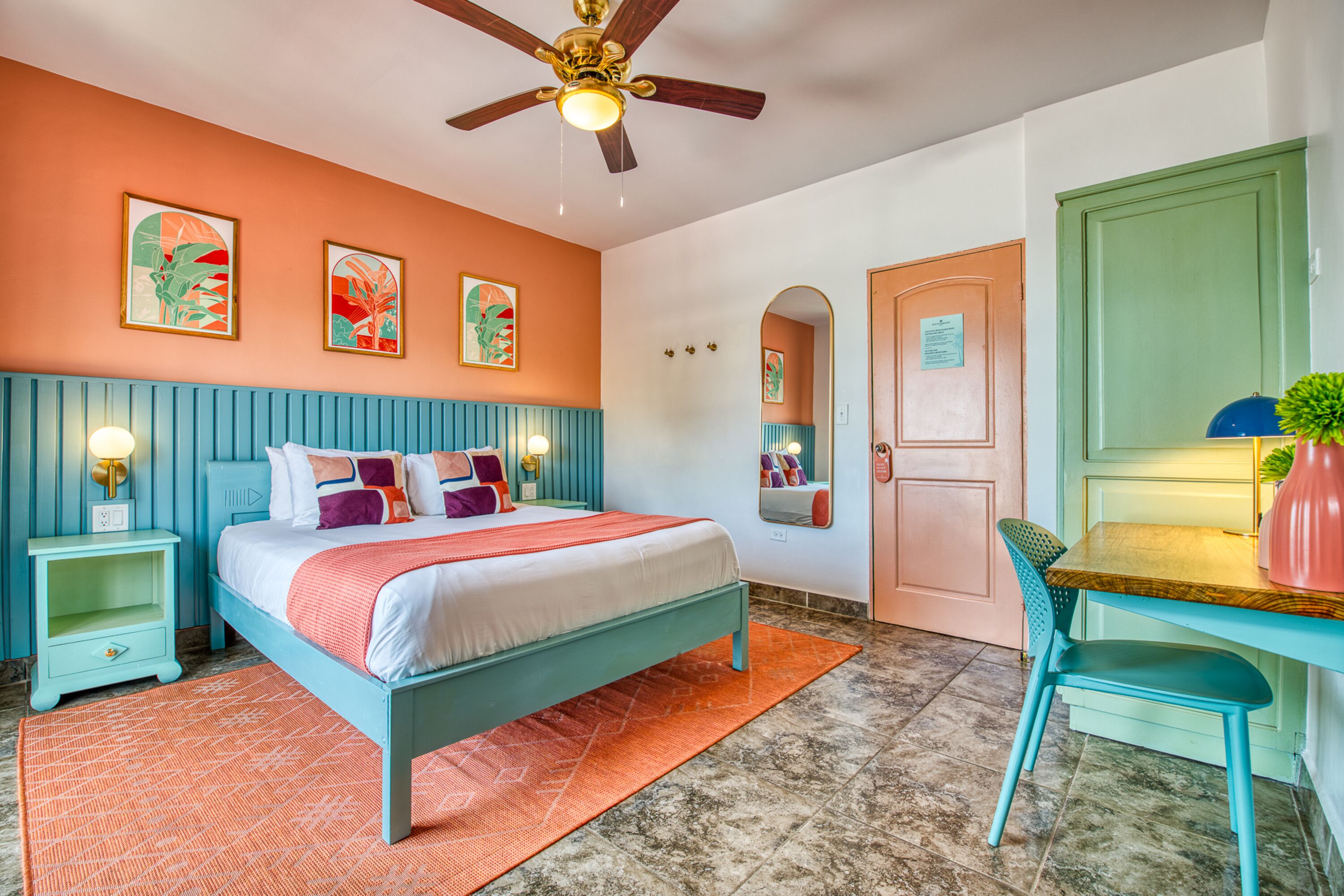 Une chambre à coucher comprenant un lit, des tables de chevet, un bureau, une chaise, un ventilateur de plafond et des œuvres d’art accrochées aux murs.