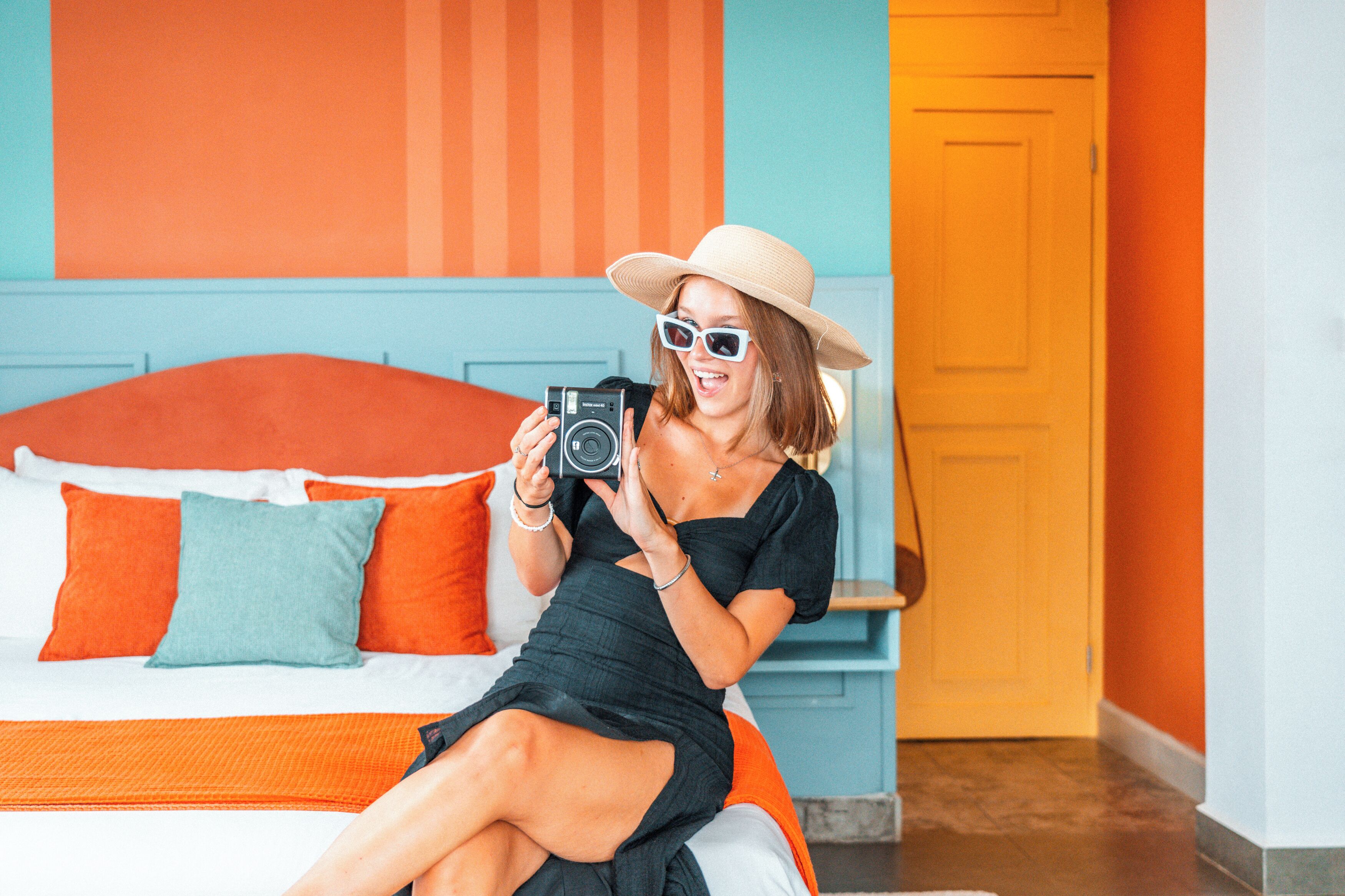 A person sitting on a bed with a camera, in a room with a blue wall and orange bedding.