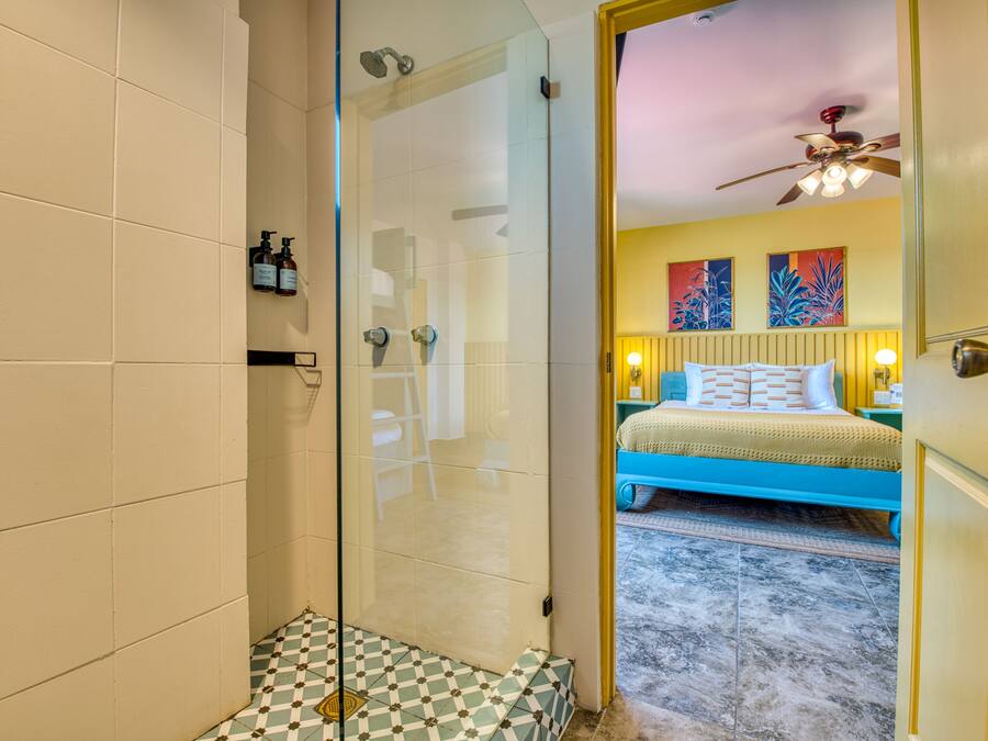 A hotel room with a glass shower enclosure, a checkered floor, a bed with blue bedding, and two paintings on the wall.