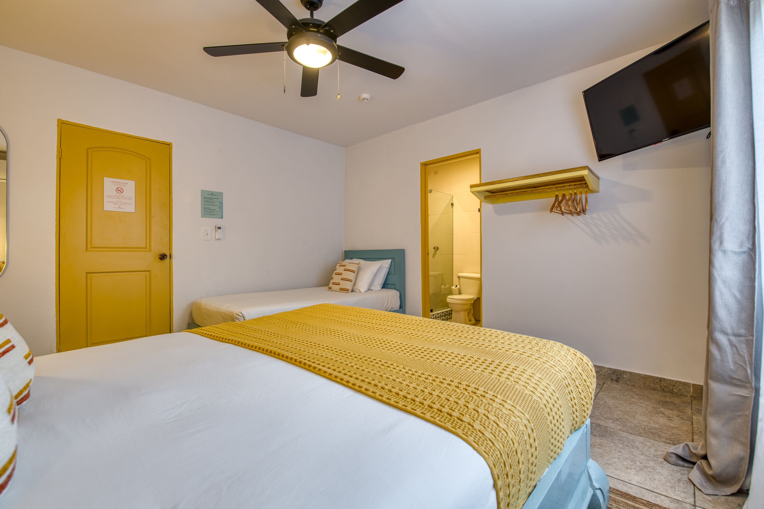 Habitación de hotel con dos camas, un ventilador de techo, un televisor montado en la pared y un baño visible a través de una puerta abierta.