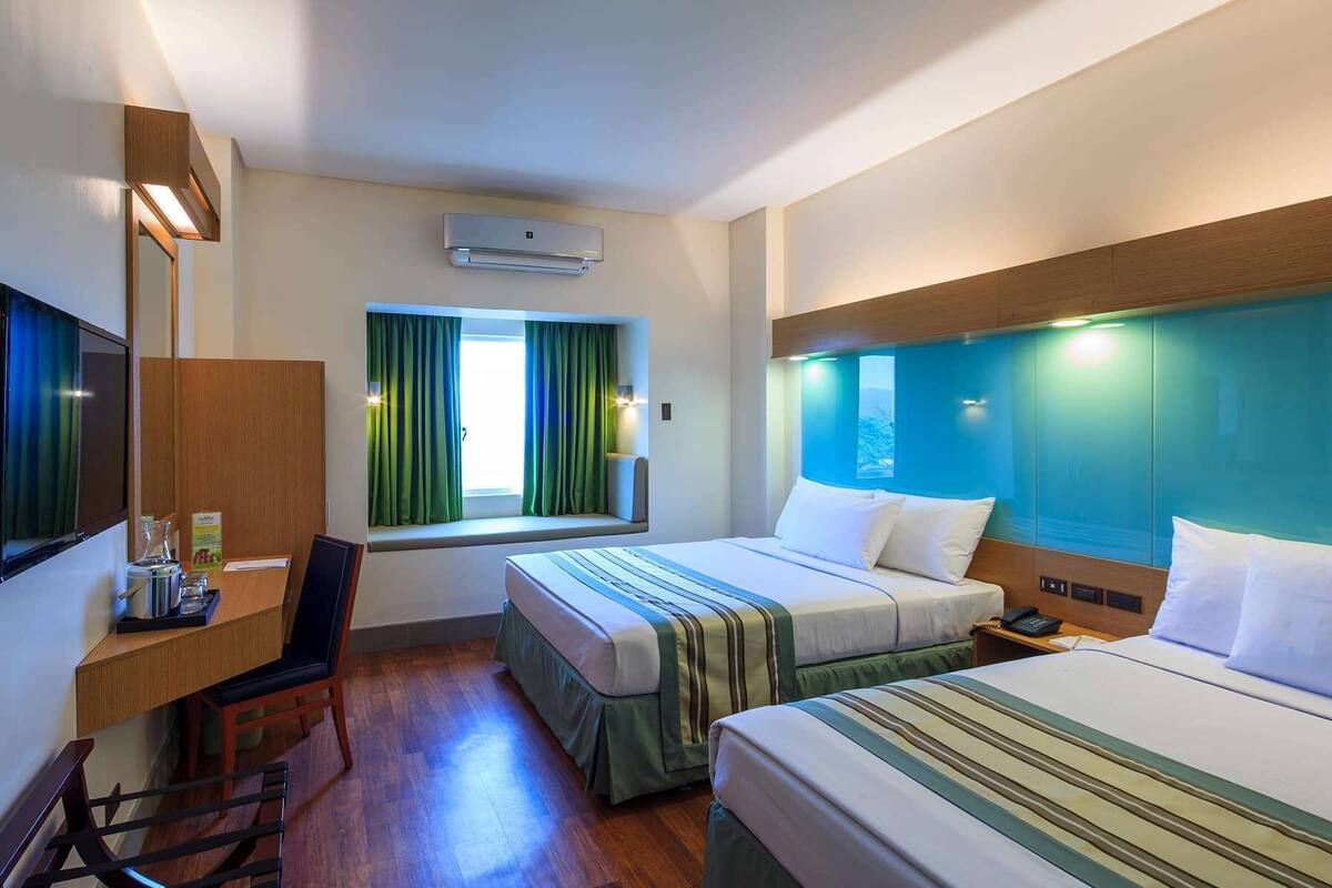 A hotel room with two beds, a desk, a chair, a window with curtains, and a mounted air conditioner.