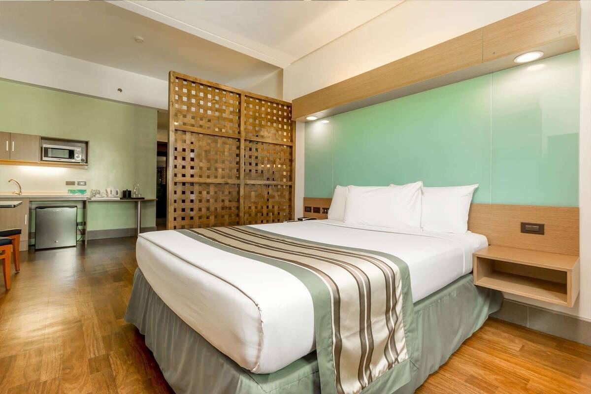 A hotel room with a bed, a wooden headboard, a kitchenette, and a partition wall.