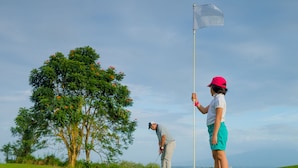 A golf course with a flagstick, a tree with orange flowers, and a clear sky.