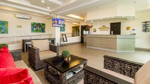 A hotel lobby with a reception desk, seating area with patterned sofas, a television screen, and artwork on the walls.