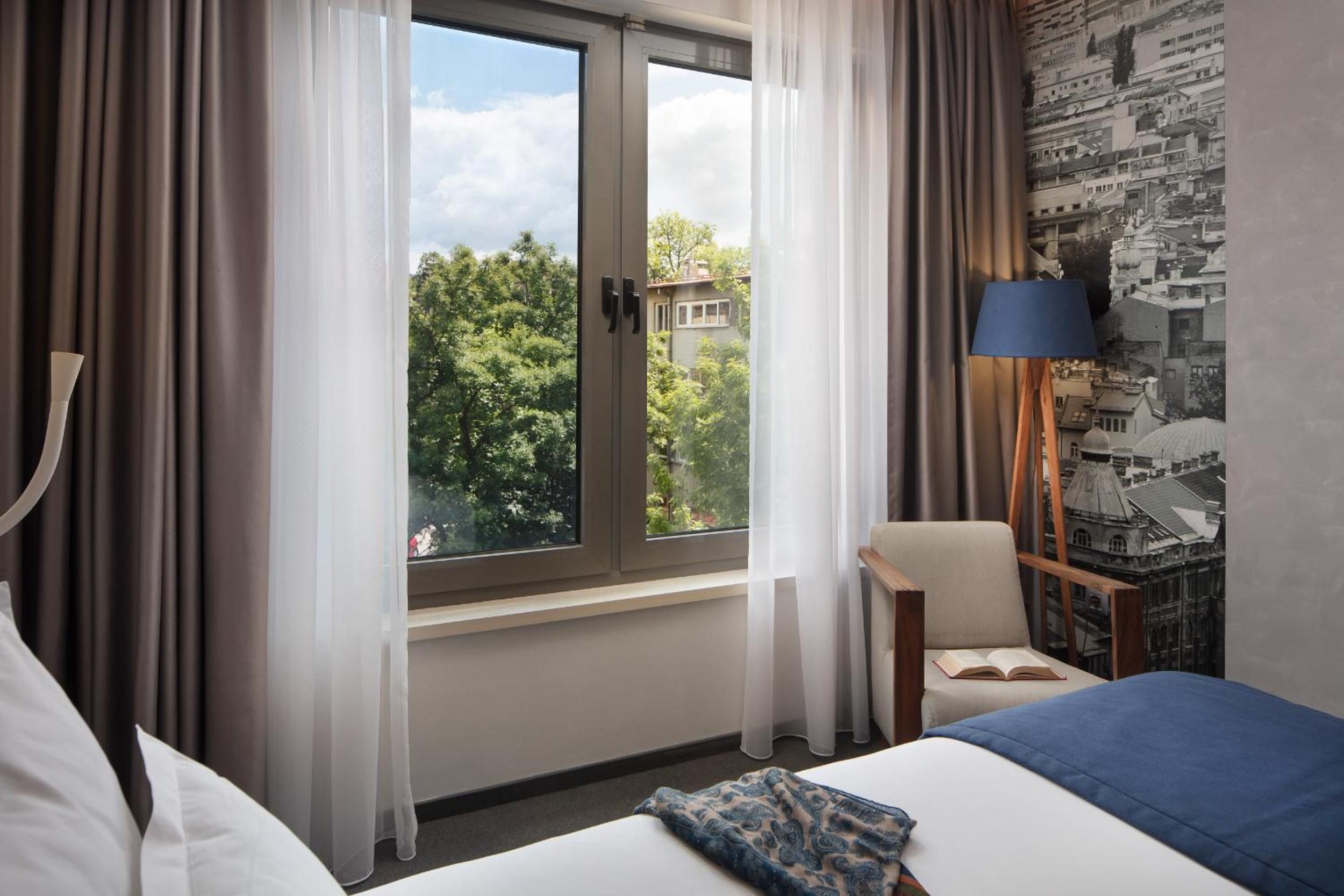 Una habitación de hotel con un ventanal grande, una cama, una silla, una lámpara y un mural urbano en la pared.