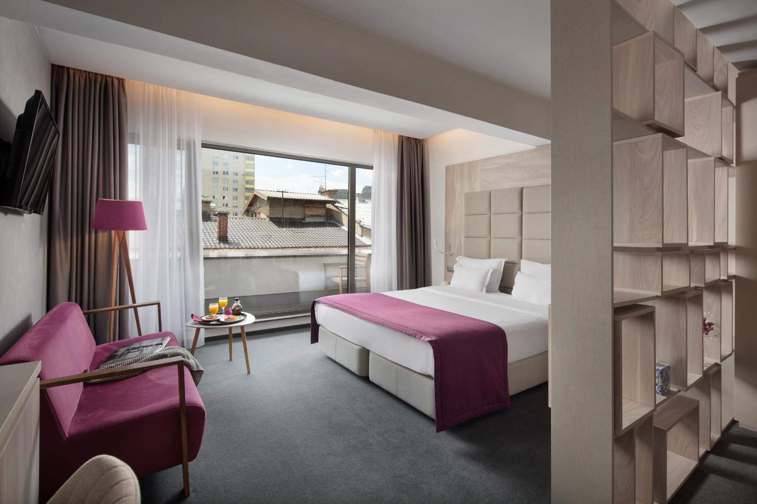 A hotel room with a bed, a desk, a chair, a small table with a lamp, and a view of buildings through a window.
