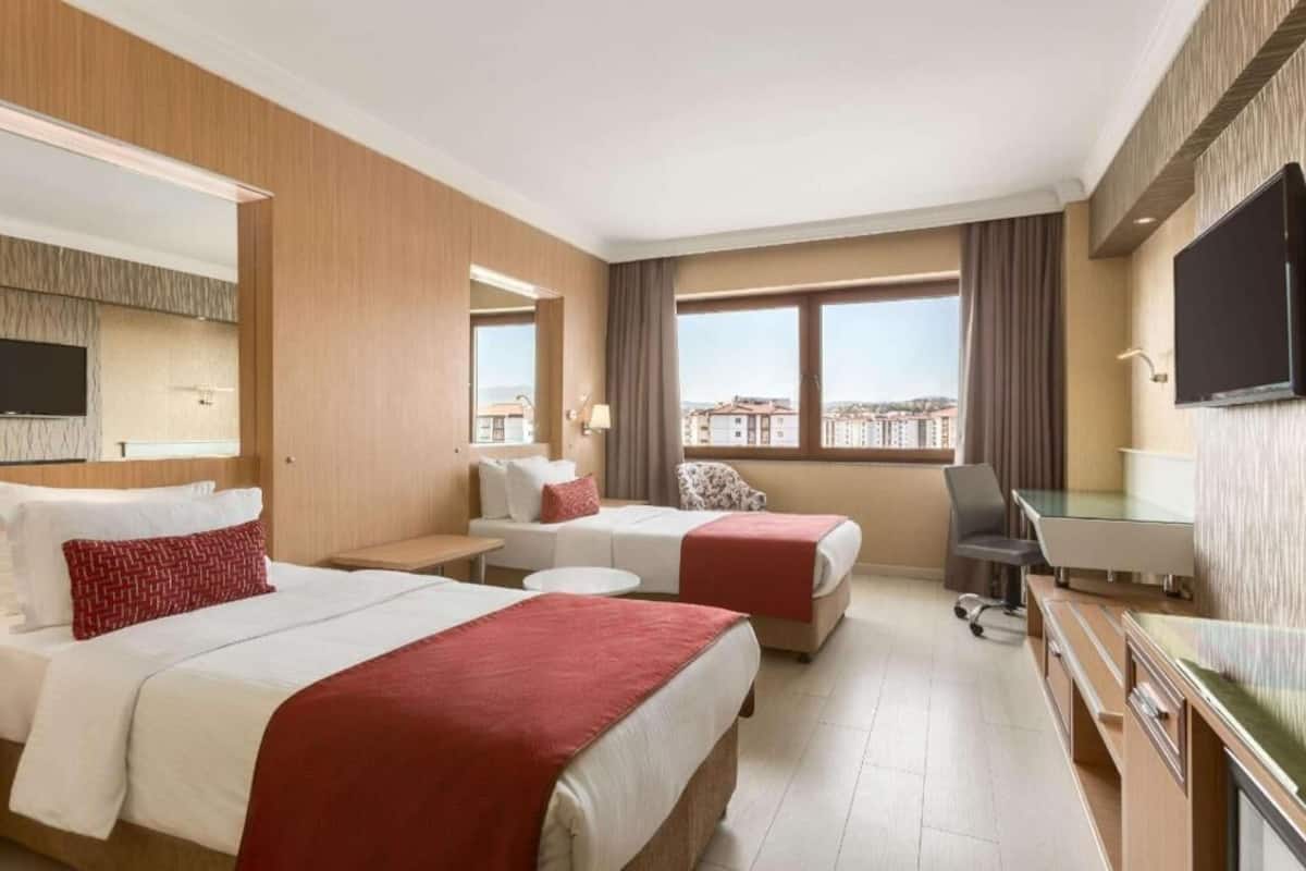 A hotel room with two beds, a desk, a chair, a TV, and a large window with a city view.