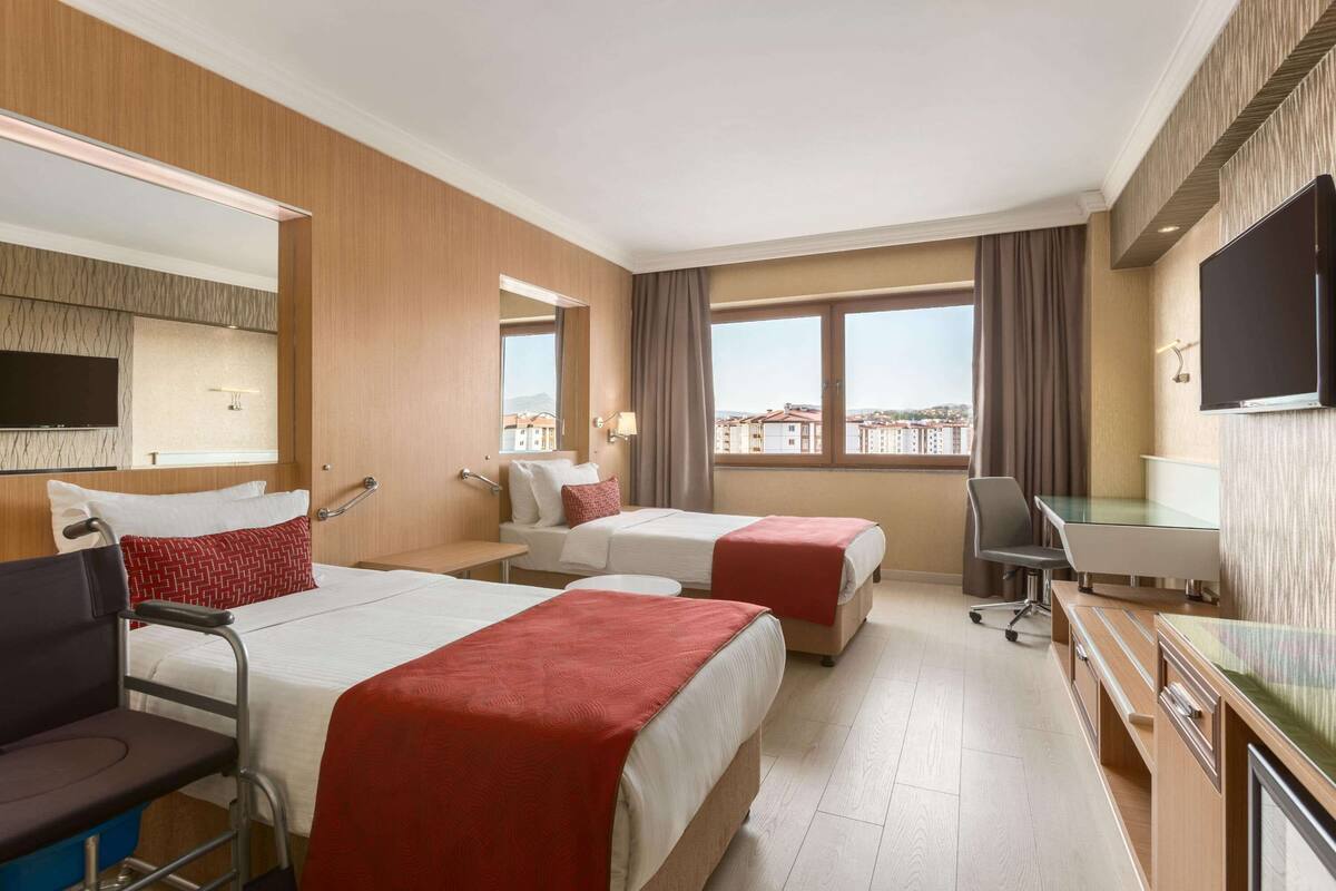 A hotel room with two beds, a desk, a chair, a TV, and a view of buildings.