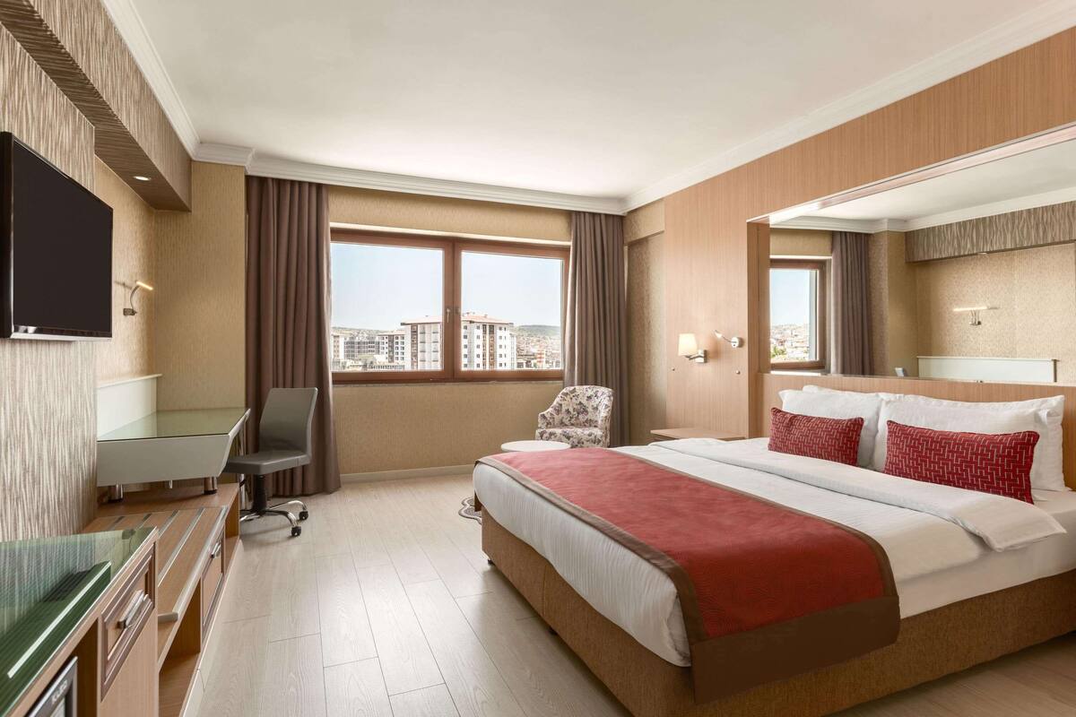 A hotel room with a large bed, a desk, a chair, a TV, and a window with a city view.