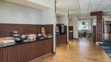 Un hall d’hôtel moderne avec un comptoir d’accueil en bois, un espace buffet proposant divers plats et un coin salon avec une table ronde et des chaises.