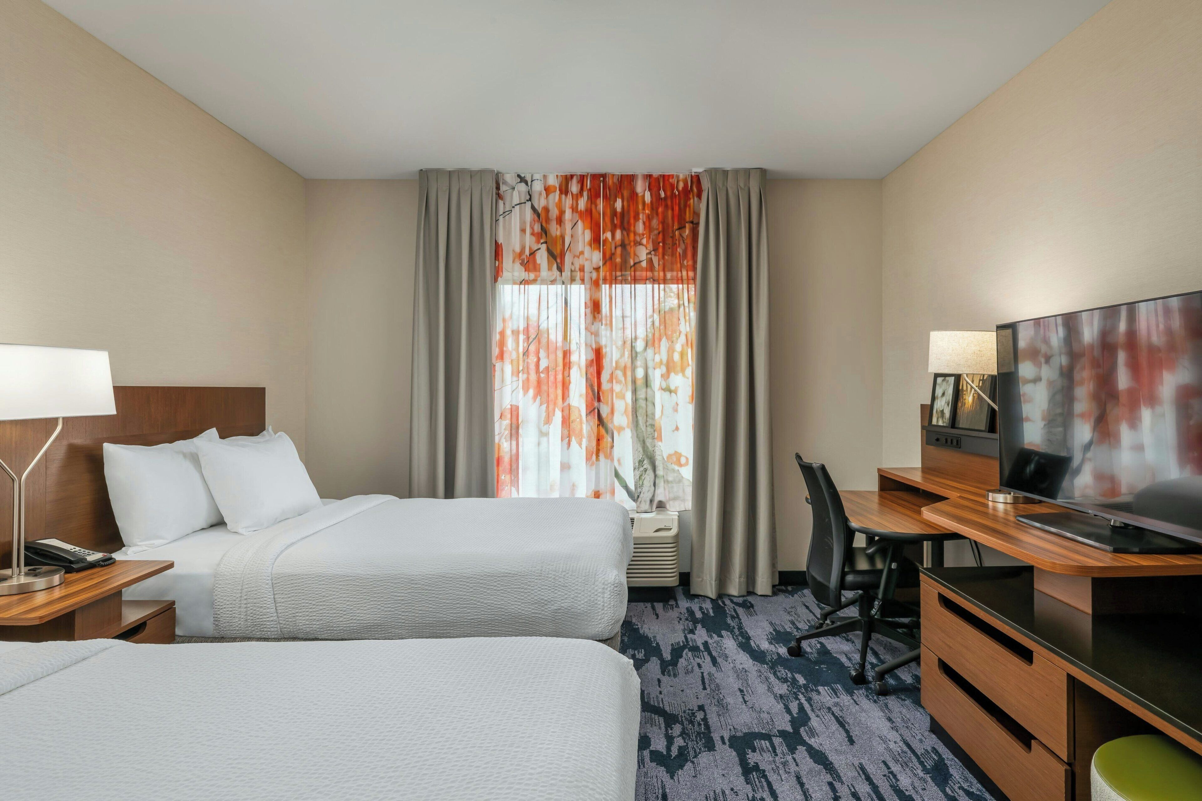 Quarto de hotel com duas camas, uma escrivaninha com televisão, uma lâmpada e uma janela com cortinas.