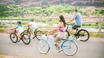 Una famiglia pedala sulle biciclette lungo un percorso asfaltato, con vista panoramica su una valle verde e scogliere sullo sfondo.
