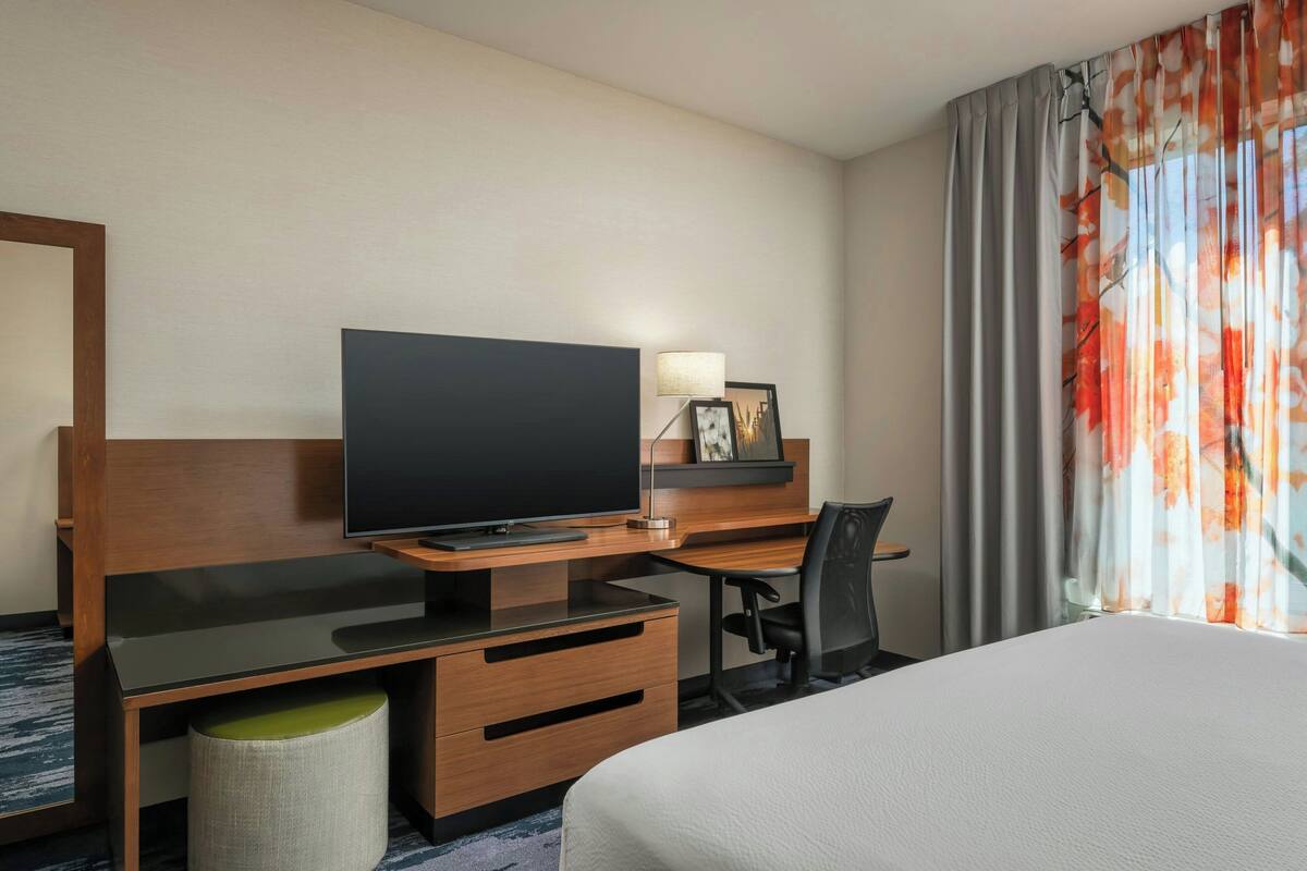 Een hotelkamer met een bed, een televisie op een houten bureau, een stoel, een lamp en een raam met gordijnen.