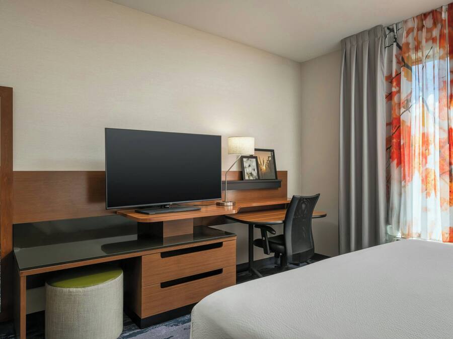 Habitación de hotel con cama, televisor sobre un escritorio de madera, una silla, una lámpara y ventana con cortinas.