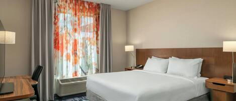 Habitación de hotel con una cama grande, un escritorio con silla, una lámpara y una ventana con cortinas.