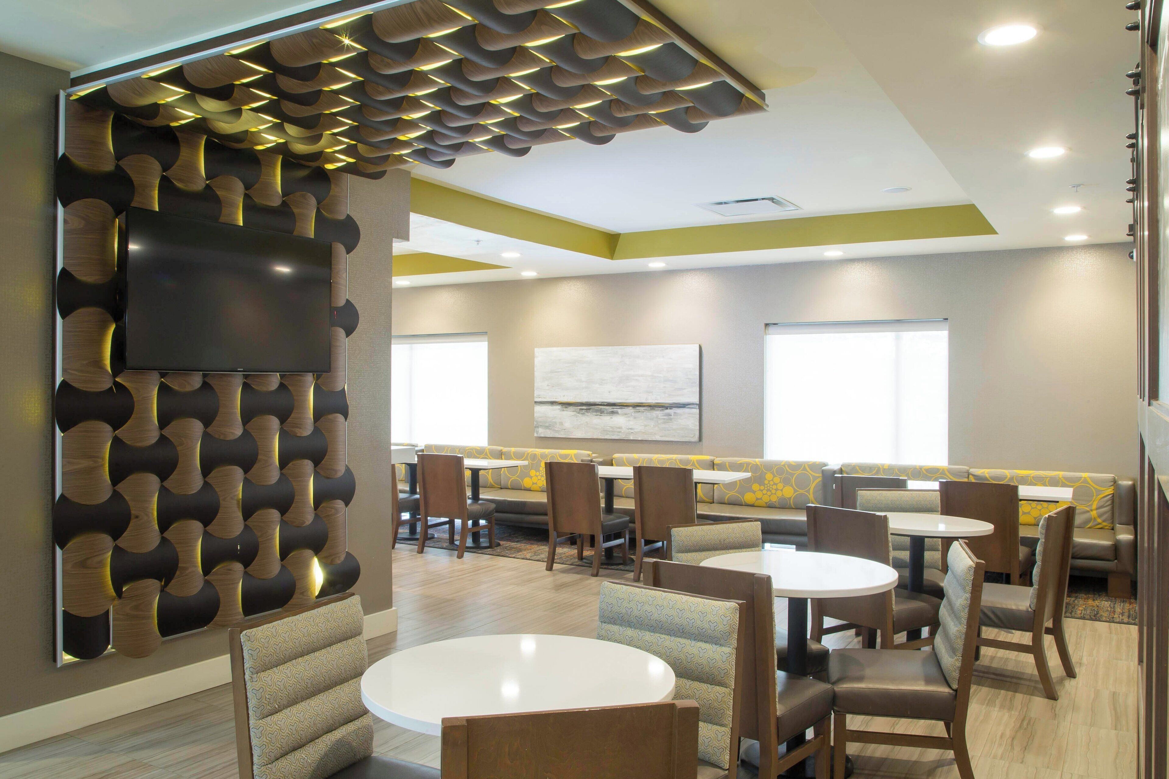 A modern dining area with round tables and booths, a flat-screen TV mounted on a textured wall, and a large abstract painting on the wall.