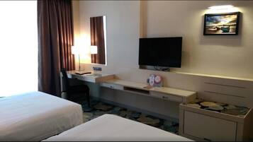 Quarto de hotel com duas camas, uma mesa de trabalho com luminária, televisão de tela plana e um pequeno armário.