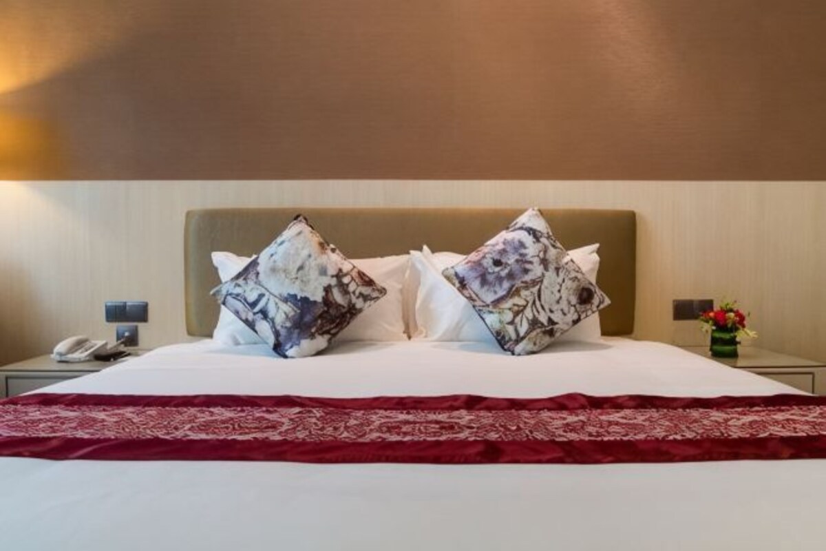 A hotel room with a bed, two decorative pillows, a red bedspread, a nightstand with a phone, and a vase with flowers.