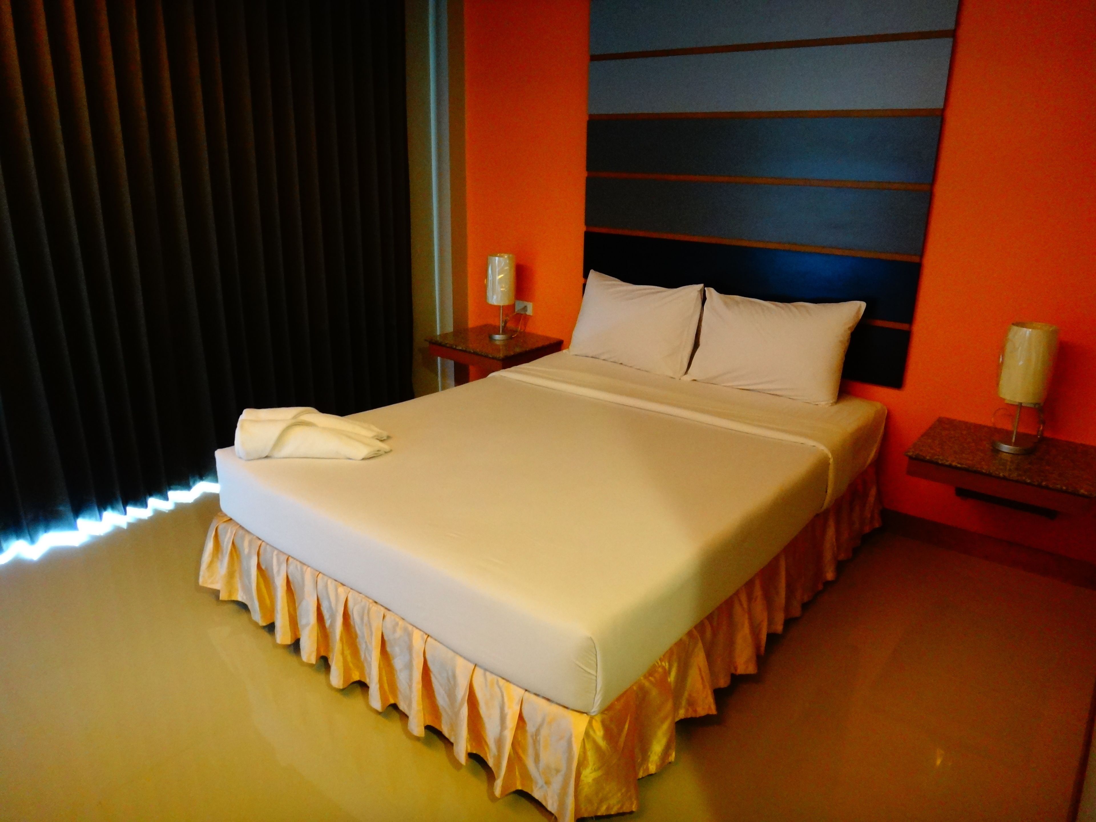 A neatly made bed with a white comforter and a decorative skirt at the foot. There is a white pillow on the bed, and a white towel is placed on the bed. The room has a modern design with orange walls and a dark window blind.