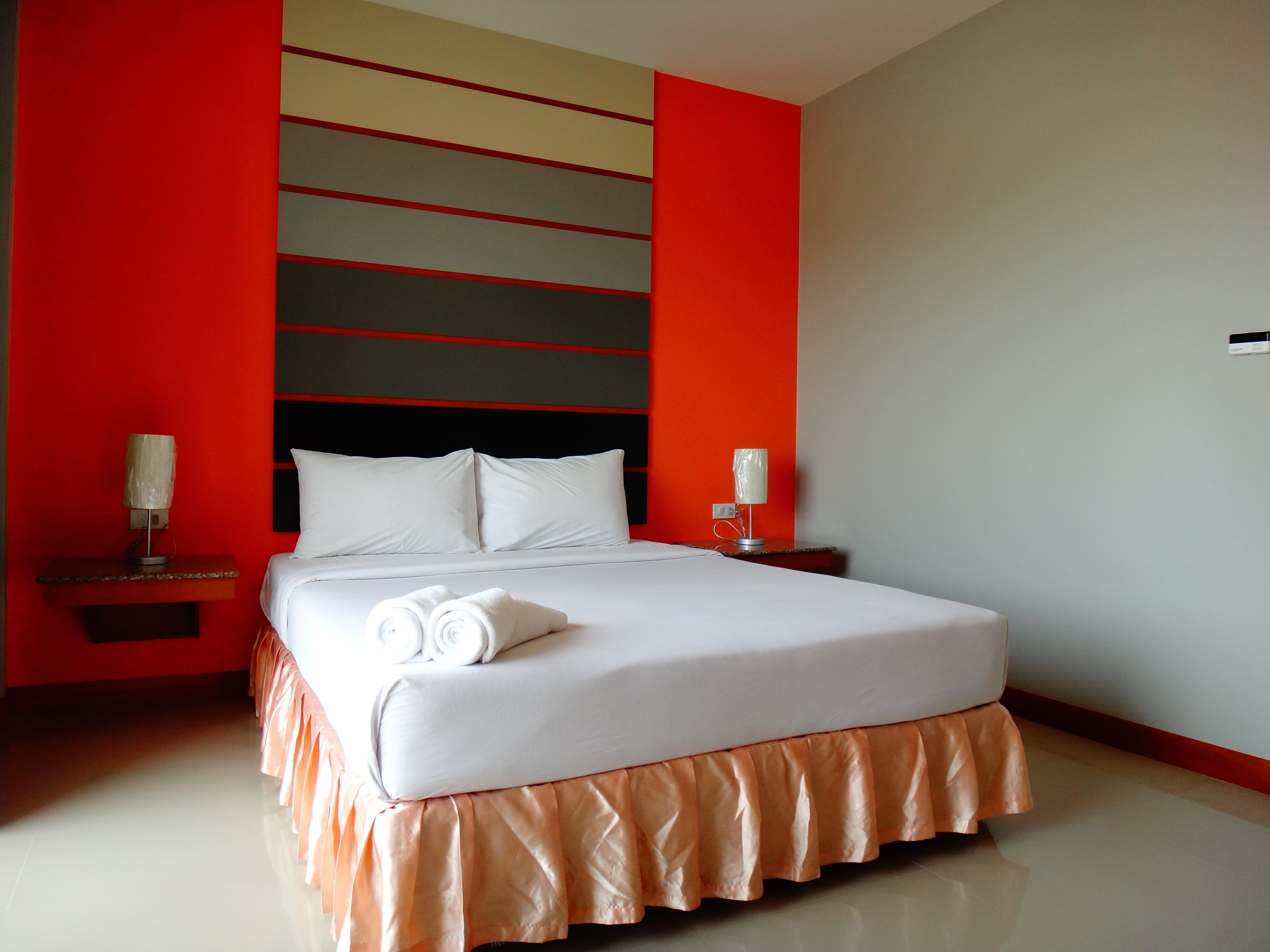 Une chambre d’hôtel avec un grand lit, du linge de lit blanc et une courtepointe de couleur pêche. Il y a deux tables de chevet avec des lampes, et les murs sont peints en rouge et en gris.