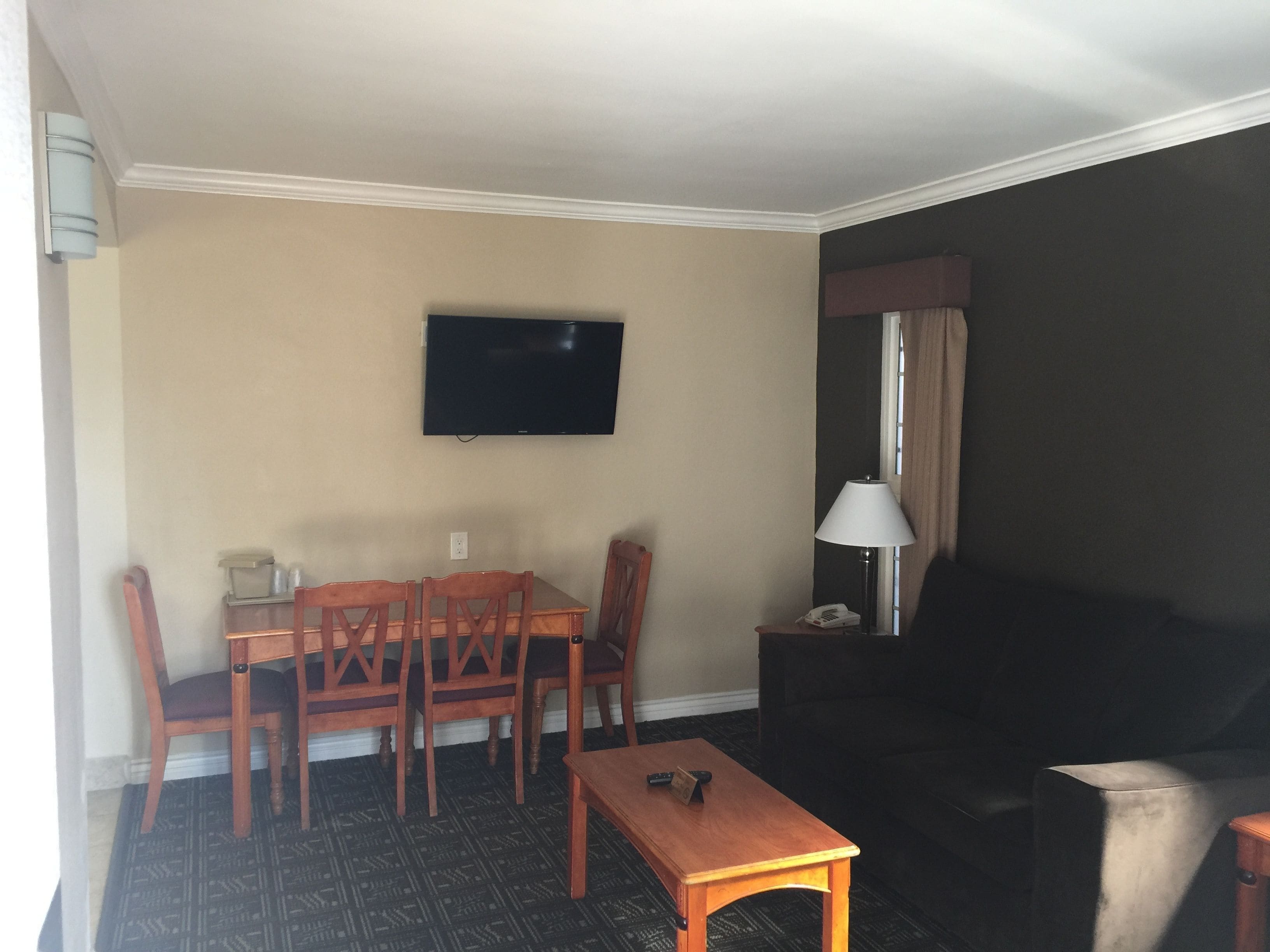 Ein Hotelzimmer mit Esstisch, Stühlen, einem an der Wand befestigten Fernseher, einer Lampe und einem Sofa.