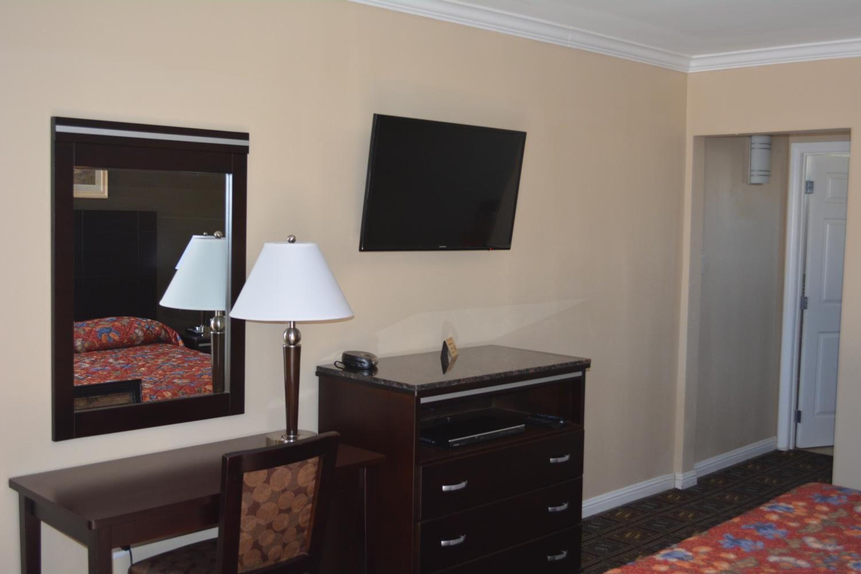 A hotel room with a bed, a desk with a lamp, a dresser, a TV mounted on the wall, and a door leading to another room.