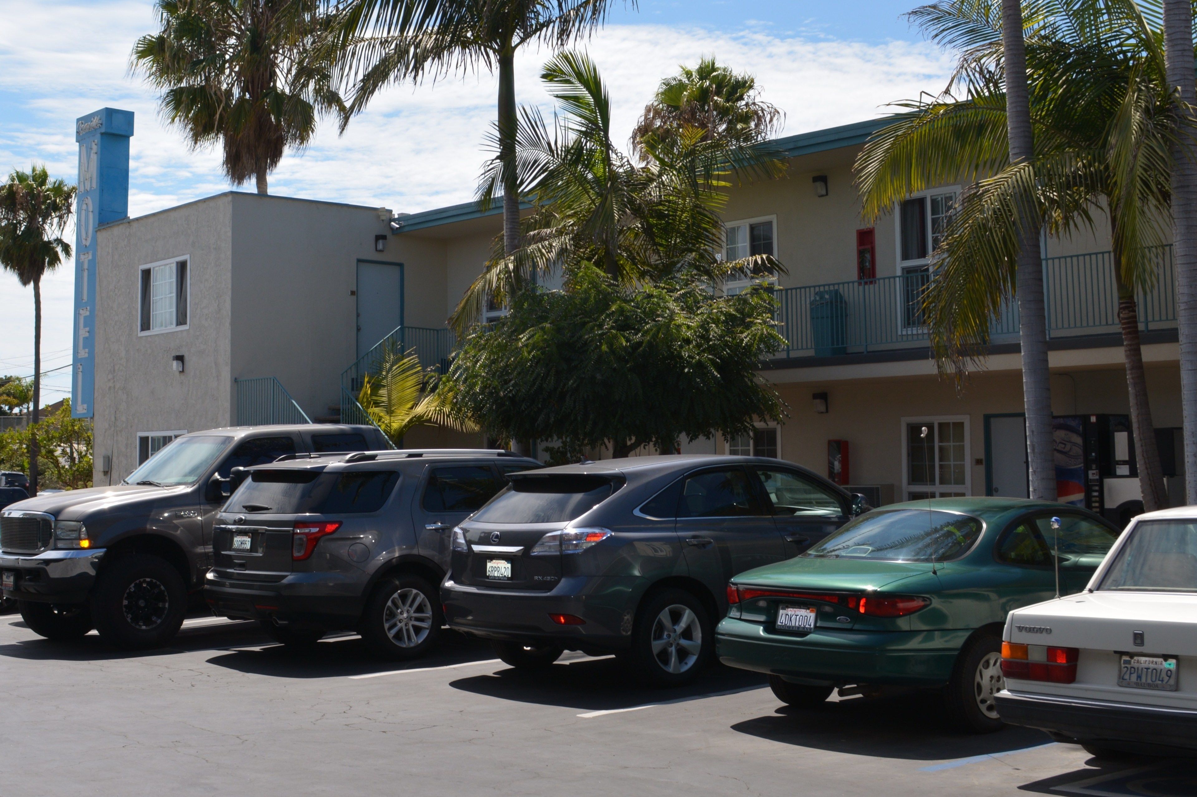 A parking lot with several SUVs and a sedan parked in front of a building with a balcony and palm trees.