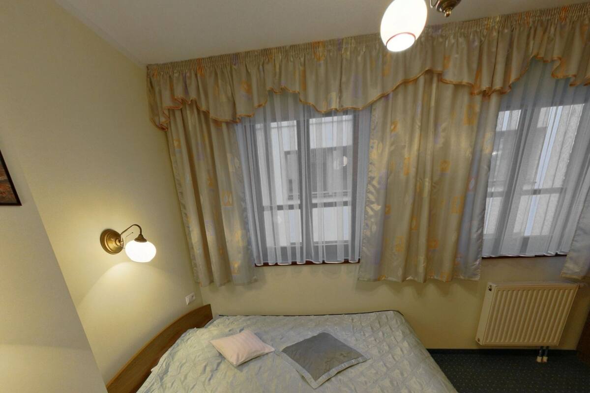 Ein Schlafzimmer mit einem Bett, einem Nachttisch, einem Fenster mit Vorhängen, einer Wandleuchte und einer Heizung.
