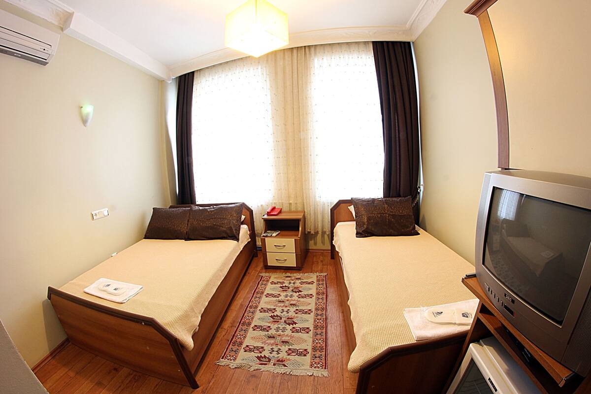 A hotel room with two single beds, a small bedside table, a TV, and a window with curtains.