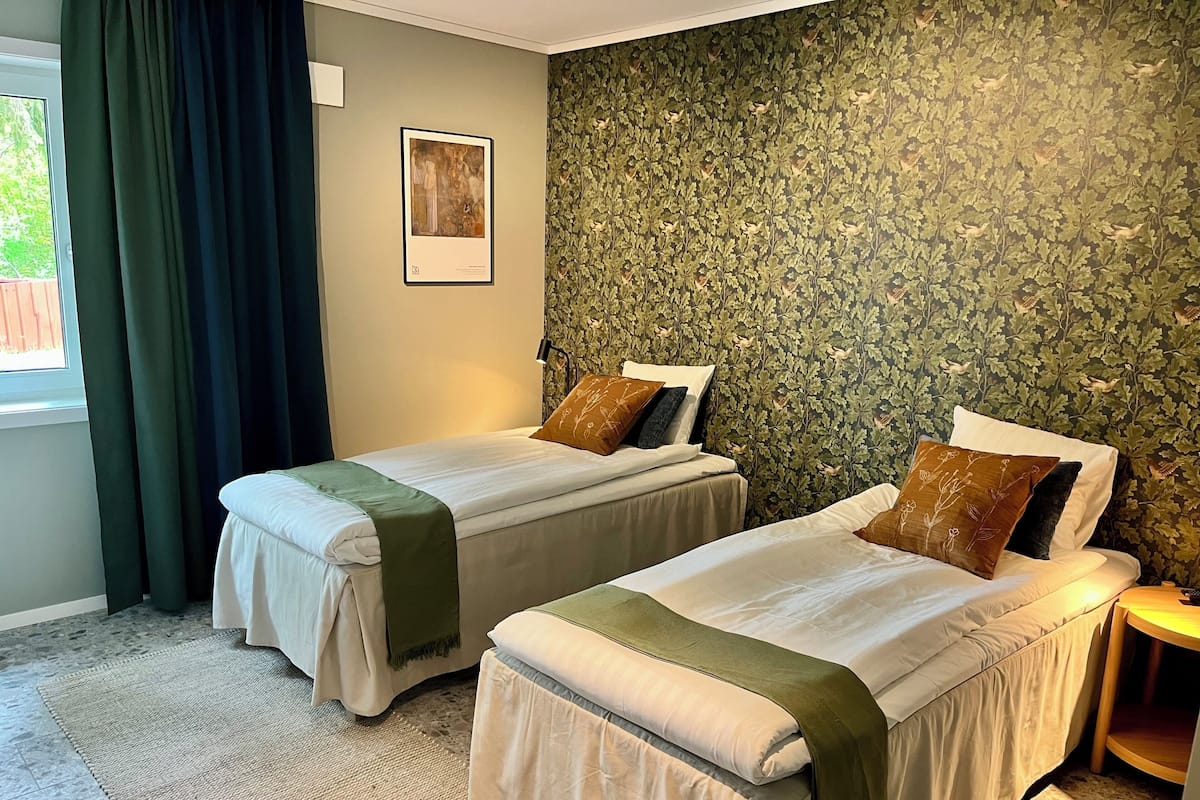 A hotel room with two beds, a green patterned wallpaper, a framed picture on the wall, and a window with curtains.