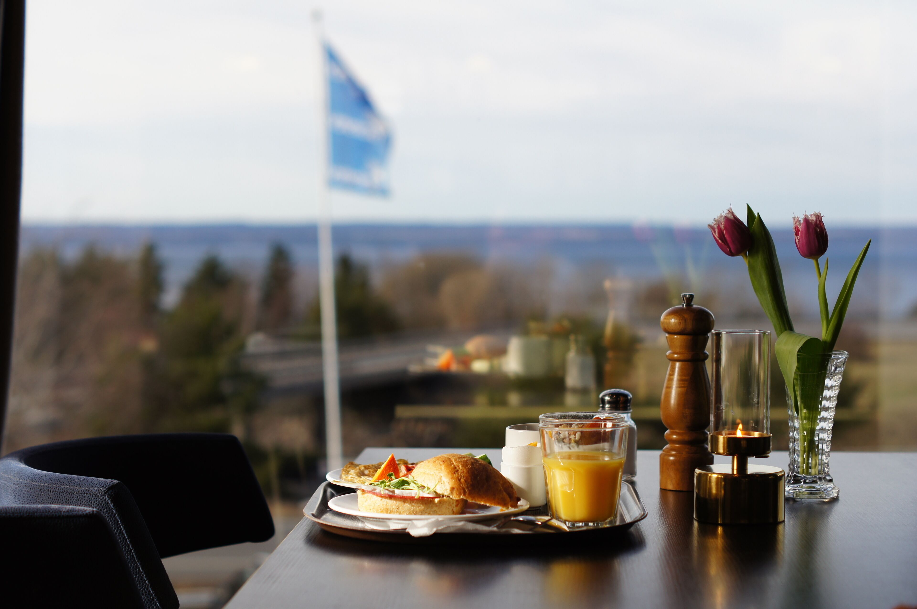 Bandeja de desayuno con un sándwich, jugo y una vela sobre una mesa con vista al mar y una bandera.