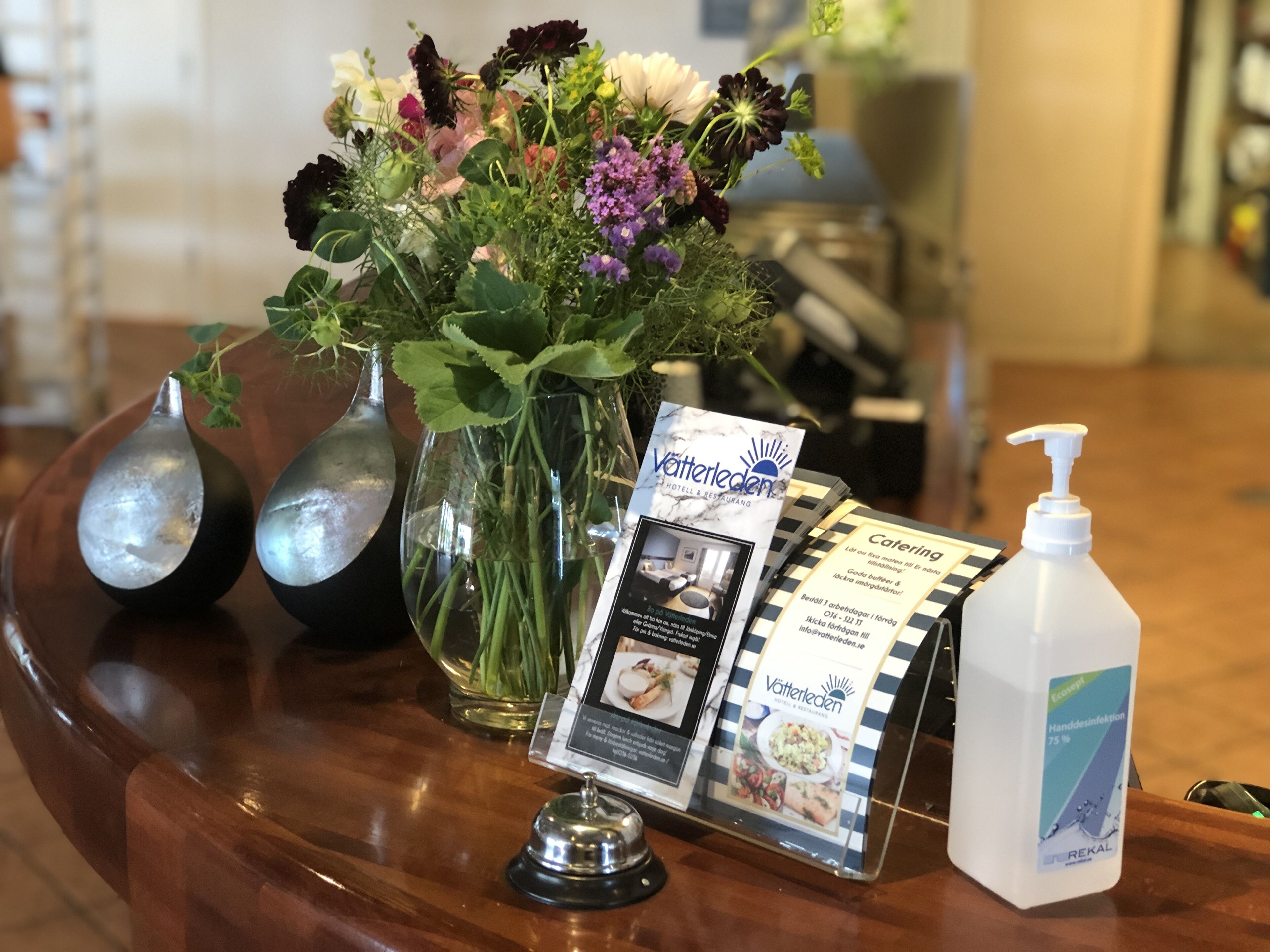 A reception desk with a floral arrangement, a brochure, a bottle of hand sanitizer, and a spoon-shaped container.