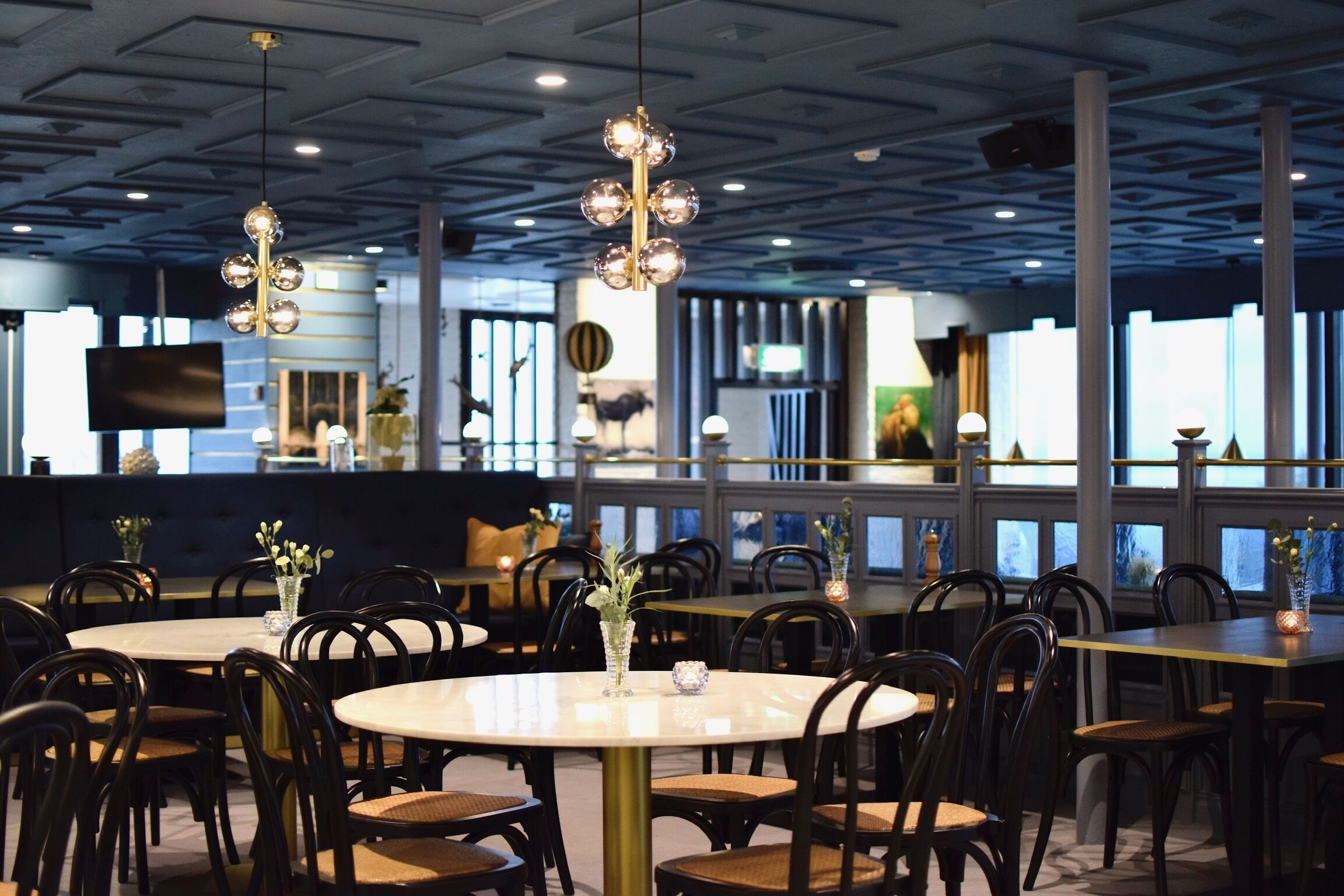 Ein Restaurant mit runden Tischen und schwarzen Stühlen, mit modernem Deckenstil und großen Fenstern.