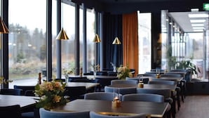 Un restaurante con mesas y sillas, ventanas amplias y lámparas colgantes.