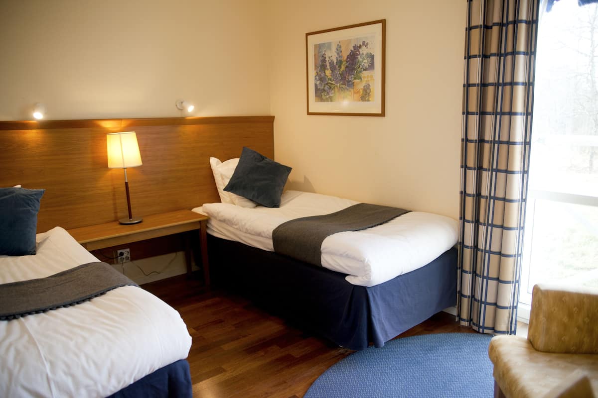 A hotel room with two beds, a bedside table with a lamp, a framed artwork on the wall, and a window with curtains.