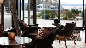 Un área de descanso con sillas y una mesa, ventanas amplias con vista a un estacionamiento y una masa de agua.