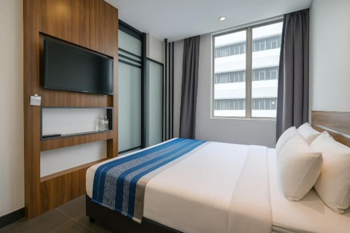 A hotel room with a bed, a TV mounted on a wooden panel, a window with curtains, and a view of a building outside.