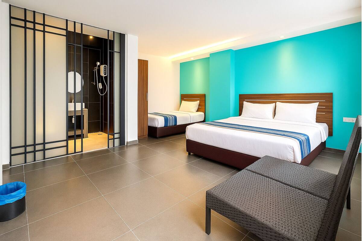 A hotel room with a large bed, two smaller beds, a bathroom with a shower, and a seating area with a chair and table.
