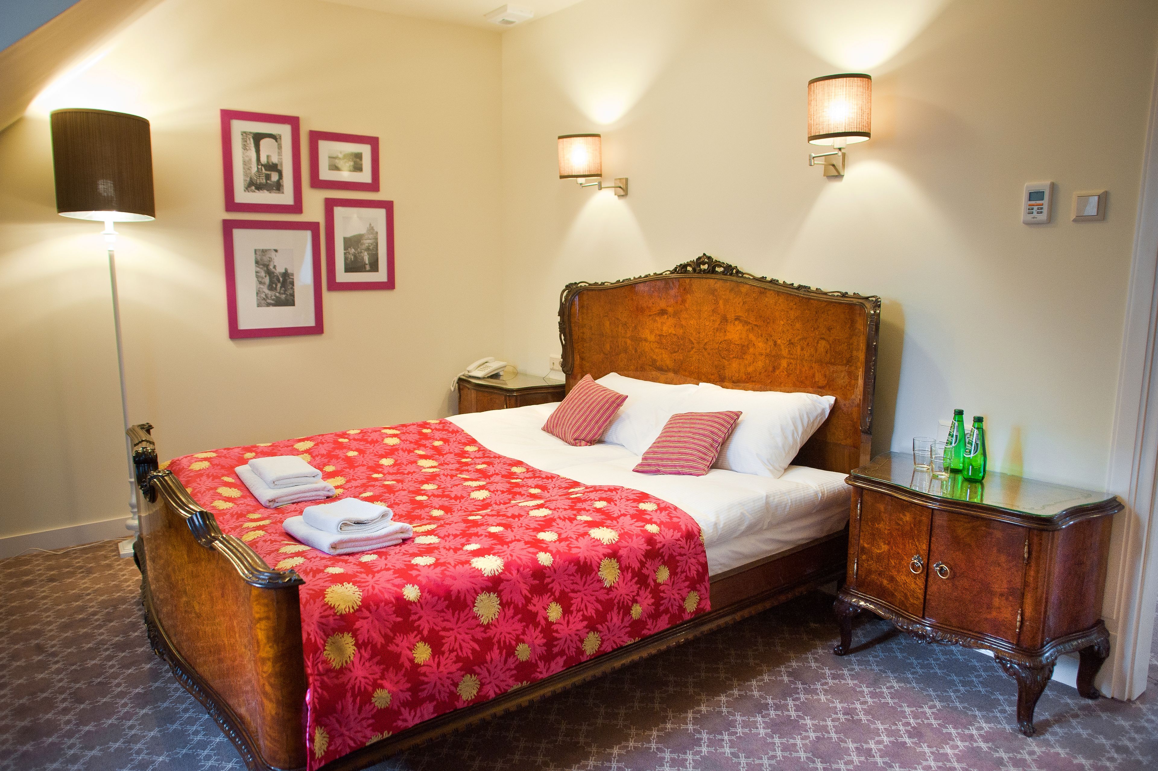 Una cama hecha con pulcritud, con una colcha floral roja, dos almohadas y toallas dobladas. Hay dos mesitas de noche, una con una lámpara y la otra con un teléfono y una botella.