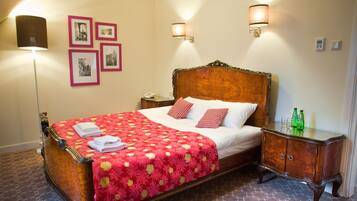 Una cama hecha con pulcritud, con una colcha floral roja, dos almohadas y toallas dobladas. Hay dos mesitas de noche, una con una lámpara y la otra con un teléfono y una botella.