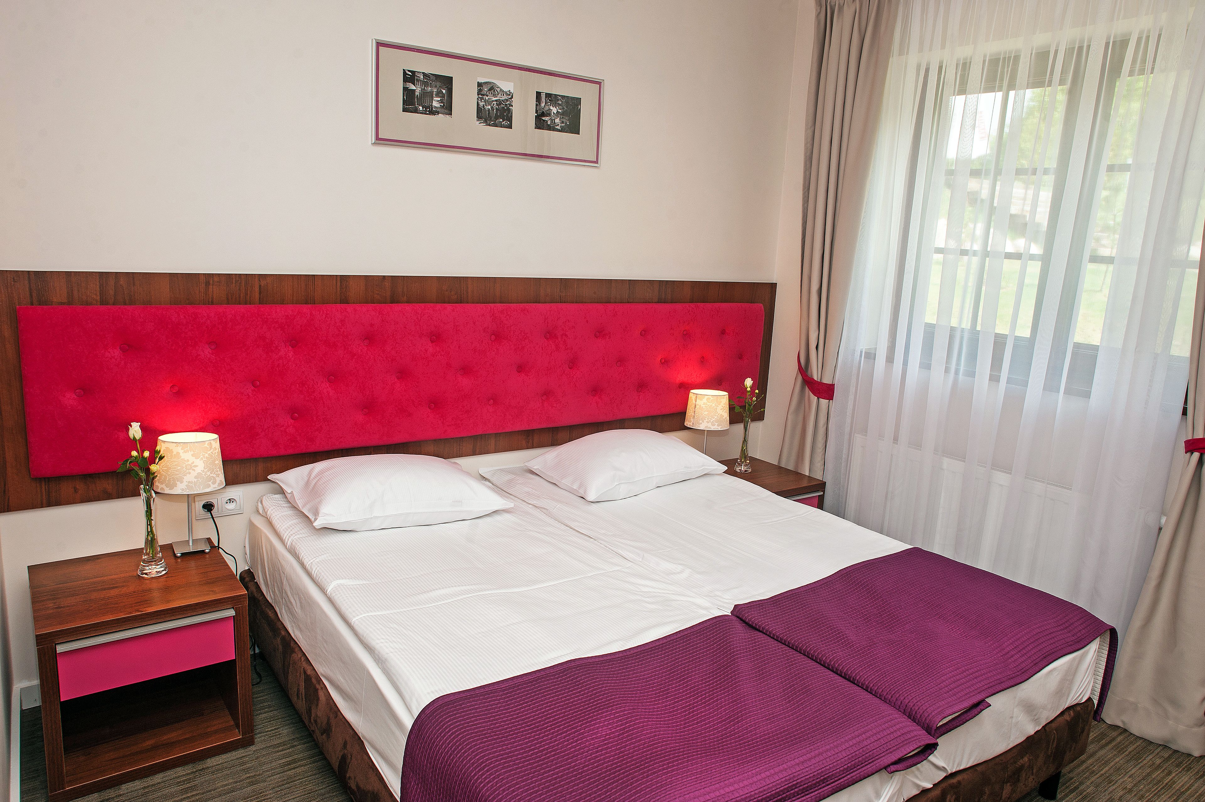 Una habitación de hotel con una cama grande, dos mesitas de noche, un cabecero rojo y una ventana con cortinas translúcidas.