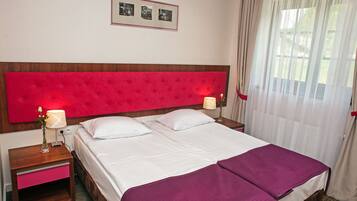 Una habitación de hotel con una cama grande, dos mesitas de noche, un cabecero rojo y una ventana con cortinas translúcidas.