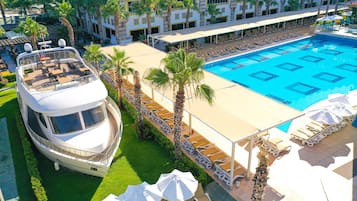 A large pool with sun loungers and umbrellas, a white boat-shaped building, and palm trees.
